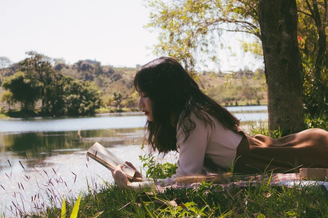 woman reading book and lying forward on sheet on grass beside body of water during day